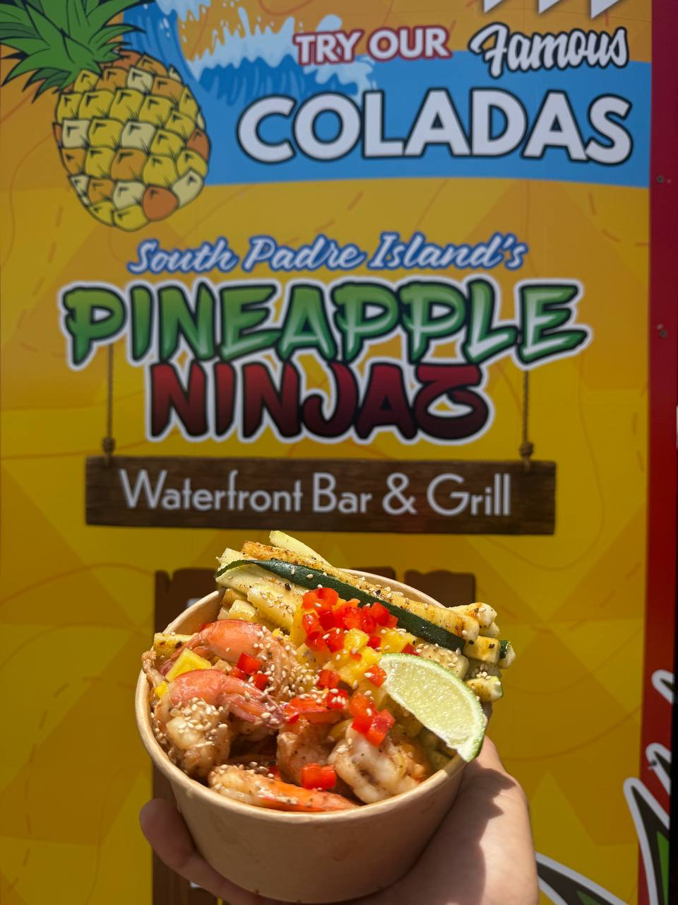 Grilled shrimp Island Bowl in a carved pineapple in front of the Pineapple Ninjaz Waterfront Bar & Grill banner